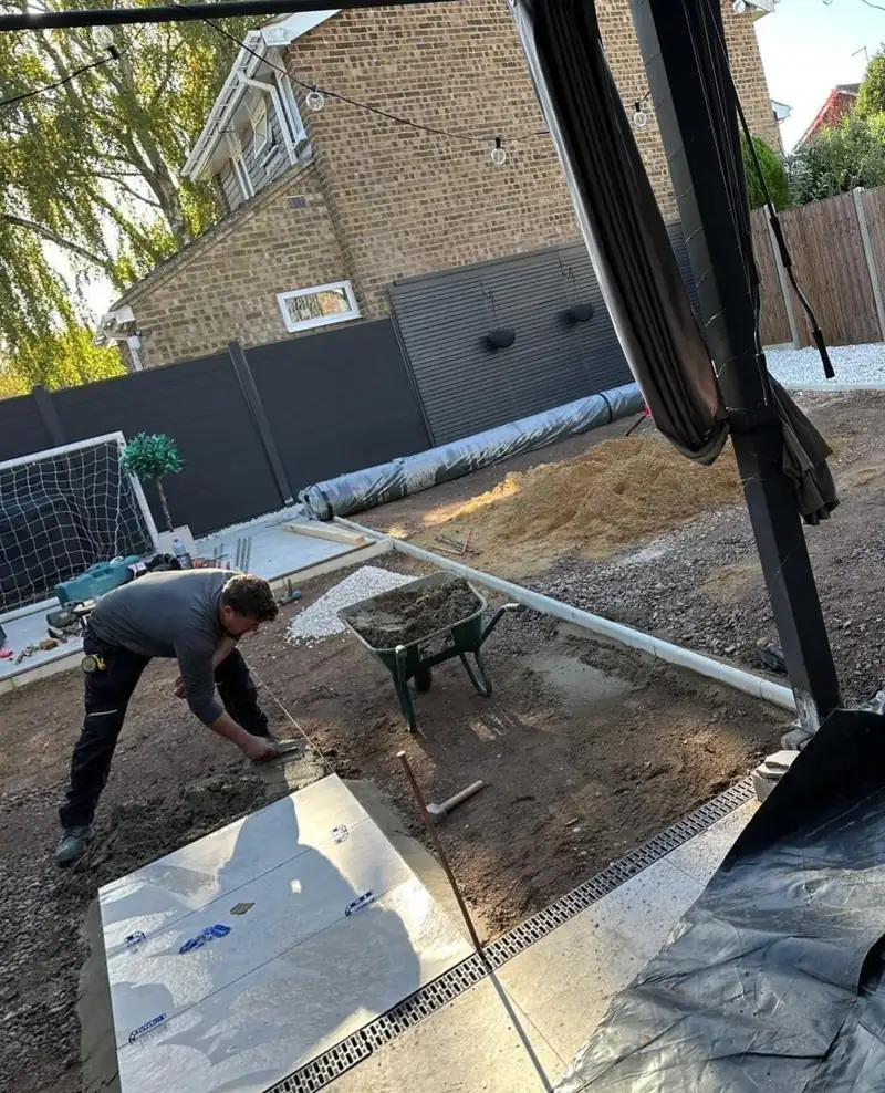A garden mid-build, with porcelain slabs being laid on a sand bed and a drainage channel along the edge