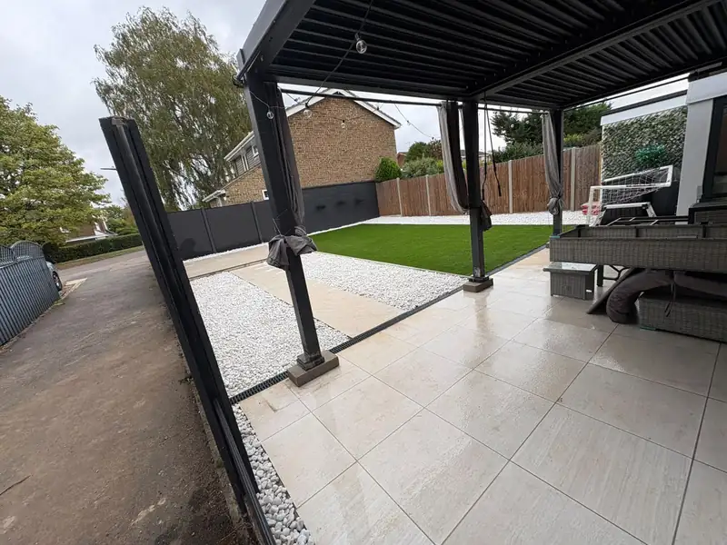 Patio under a pergola looking across white gravel and a lawn to the garden fence